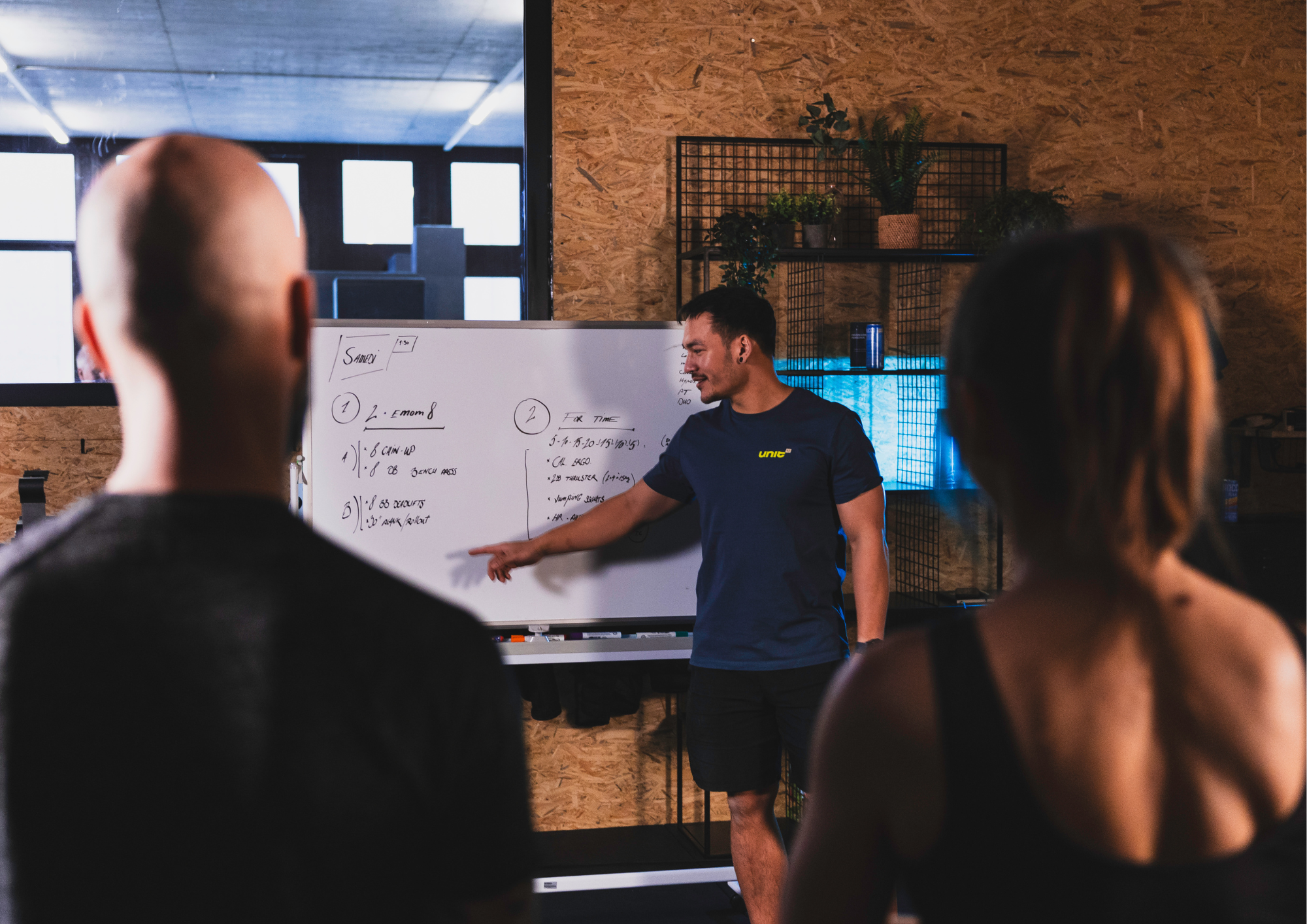 A professional and dynamic fitness coach correcting the posture of a 40-year-old participant doing a deadlift in a functional training gym equipped with kettlebells and racks. No identifiable faces.