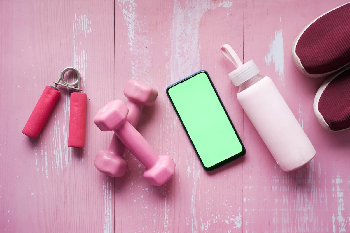 Haltères roses, smartphone à écran vert, bouteille blanche, tapis de yoga sur fond rose.