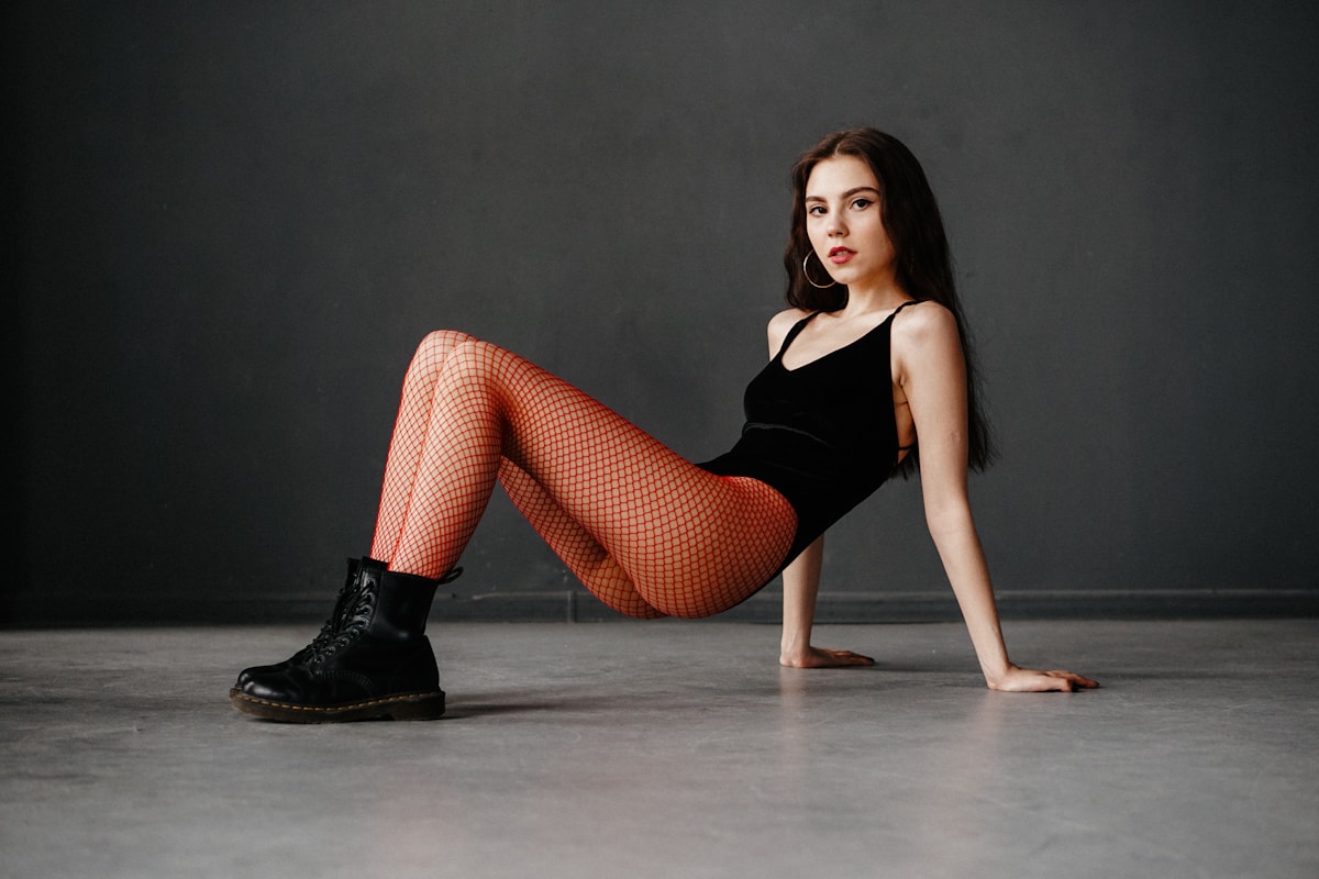 Jeune femme en tenue noire et bas résille, posant sur un sol sombre.