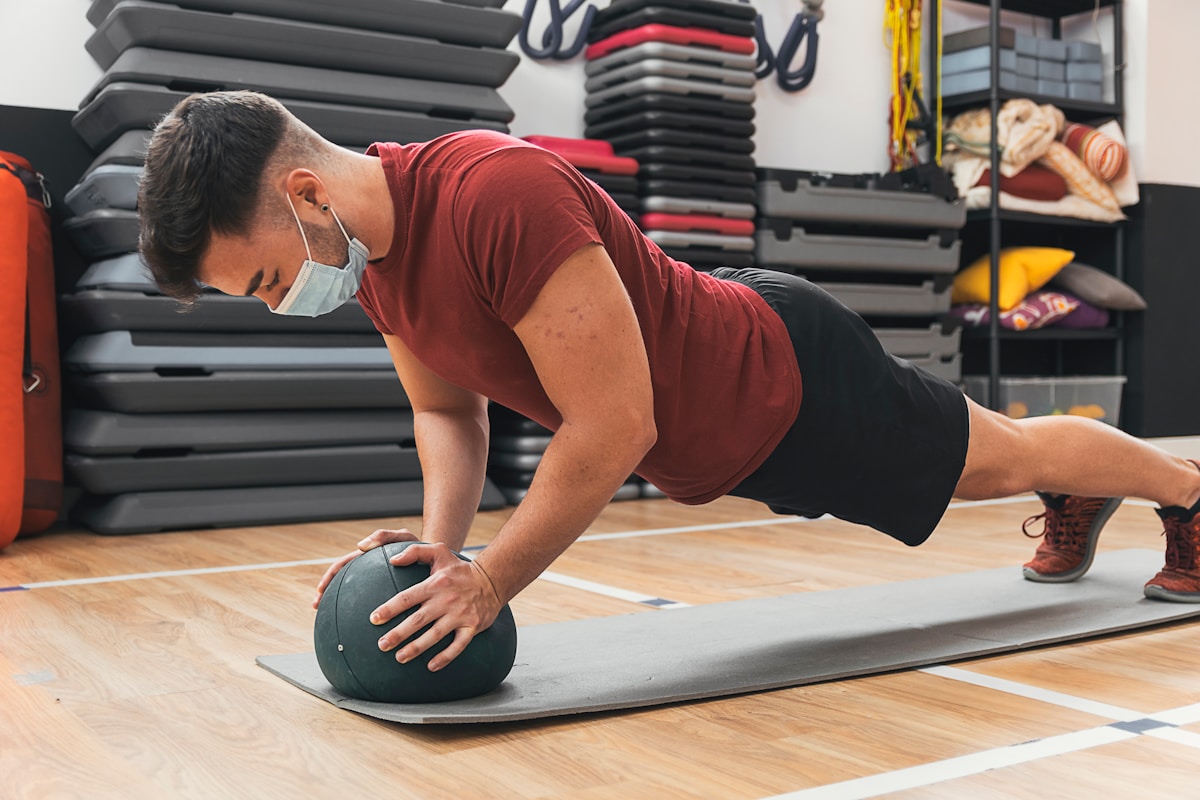 Homme en tenue de sport effectuant un exercice de planche avec un ballon médicinal.